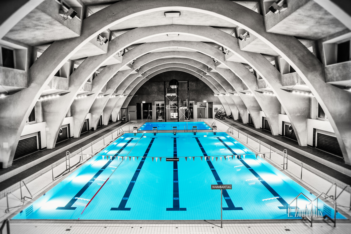 Fotografie im Inneren des Heslacher Hallenbads in Stuttgart. Schwarzweiss mit blauem Wasser in dem sich die wunderschöne Architektur spiegelt.