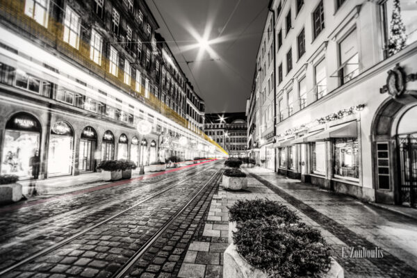 Schwarzweiss-Aufnahme in der Perusastraße in München. Farbige Lichtschweife in Gelb und Rot entlang der Schienen