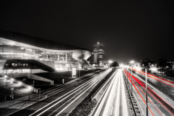 Schwarzweiss-Aufnahme an der BMW World in München. Im Hintergrund das Museum und das Gebäude in der Form des Vierzylinders. Rote Lichtschweife, die den Verkehr kennzeichnen