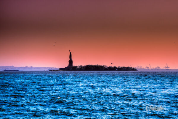 Aufnahme an der Freiheitsstatue in New York. Im Vordergrund blaues Wasser, in der Mitte des Bildes die Freiheitsstatue und im Hintergrund Industrieanlagen und Hubschrauber