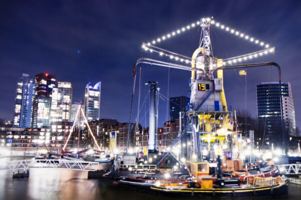 Nachtaufnahme am Hafen von Rotterdam. Im Vordergrund Schiffe auf der Anlegestelle und im Hintergrund die Hochhäuser mit funkelnden Lichtern