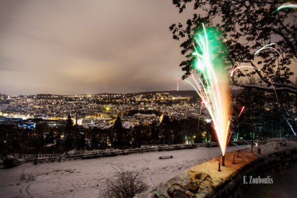 Nachtaufnahme an der Karlshöhe in Stuttgart. Im Vordergrund ein Feuerwerkskörper. Im Hintergrund Schnee und der Blick auf die Stuttgarter Innenstadt und den Fernsehturm