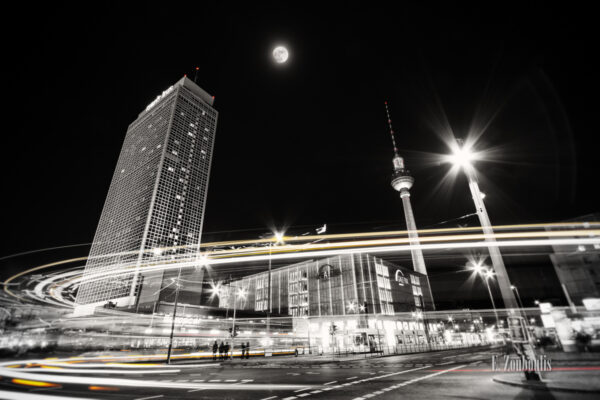 Schwarzweiß-Aufnahme am Alexanderplatz in Berlin. Die Dynamik der Stadt bei Nacht ist in der Form eines gelb / roten Lichtschweifs zu sehen, der um die Ecke schießt. Im Hintergrund erkennt man den Fernsehturm und das Park Inn Hotel bei Vollmond