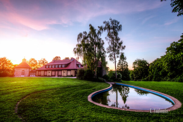 Aufnahme bei Sonnenuntergang an der Villa Schwalbenhof in Gärtringen. Im Vordergrund ist der Teich und die Wiese zu sehen und links im Bild die Villa an der sich so langsam die Sonne herablässt