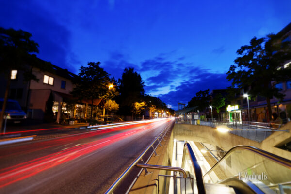 Abendaufnahme in der Kirchheimer Straße in Stuttgart Sillenbuch. Links im Bild der Verkehr in Form von rot / orangenen Lichtschweifen. Rechts geht es durch U-Bahn Haltestelle hinab