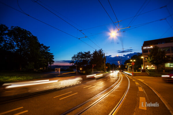 Abend-Aufnahme zur Blauen Stunde am Bubenbad in Stuttgart. Im Vordergrund sieht man die Straßenbahn-Schienen und die Fahrzeuge, die am Betrachter vorbeifahren. Links in der Mitte ist schemenhaft die Stuttgarter Innenstadt zu sehen