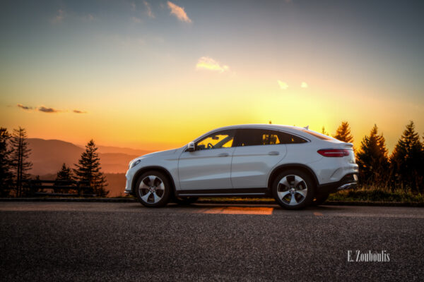 Sonnenuntergang im Schwarzwald. Die Sonne steht tief hinter einem Mercedes GLE. Seitlich sind die Nadelbäume und links im Bild die Silhouette der Berge zu sehen