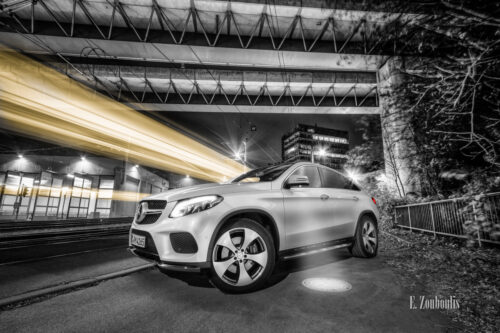 Schwarzweiß Aufnahme mit einem gelben Lichtschweif, der eine vorbeifahrende Straßenbahn kennzeichnet und roetn Elementen der Heckleuchten eines weißen Mercedes GLE unter einer Brücke in Stuttgart Südheim. Bild entstanden auf Einladung von Mercedes Benz