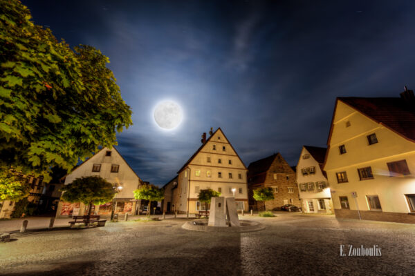 Nachtaufnahme am Marktplatz in Gärtringen bei Vollmond. Im Vordergrund der Pflasterstein und die Blätter des Ahorn Baumes. In der Mitte des Bildes der Brunnen am Marktplatz und im Hintergrund der Vollmond, der dich Dächer der Häuser erhellt