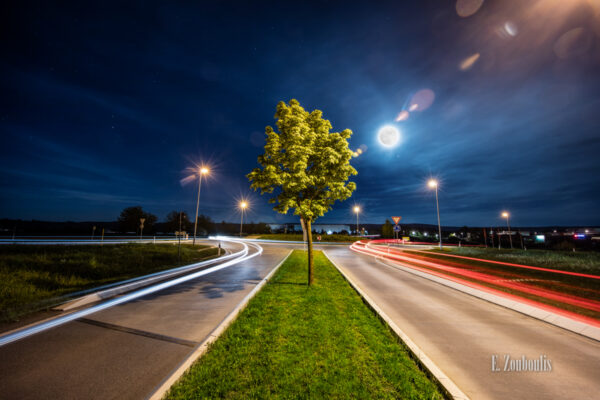 Nachtaufnahme in Gärtringen bei Vollmond. Im Vordergrund ein Baum und die Lichtschweife des Verkehrs, die zum Kreisverkehr hin- und vom Kreisverkehr wegfahren. Im Himmel der Vollmond, der die Wolken erhellt und bläulich schimmern lässt