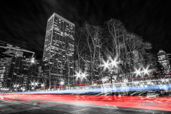 Schwarzweiß Aufnahme nachts in New York City mit einem gelben und blauen Lichtschweif, der den Verkehr kennzeichnet. Im Hintergrund Bäume und die Wolken, die über den Spitzen der Hochhäuser zu sehen sind