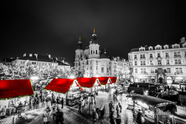 Schwarzweiß Aufnahme am Weihnachtsmarkt in Prag. Im Vordergrund sind schemenhaft die Besucher zur Weihnachtszeit zu sehen. In der Mitte des Bildes rote Dächer der kleinen Verkaufsstände und im Hintergrund die die St. Nikolaus Kirche