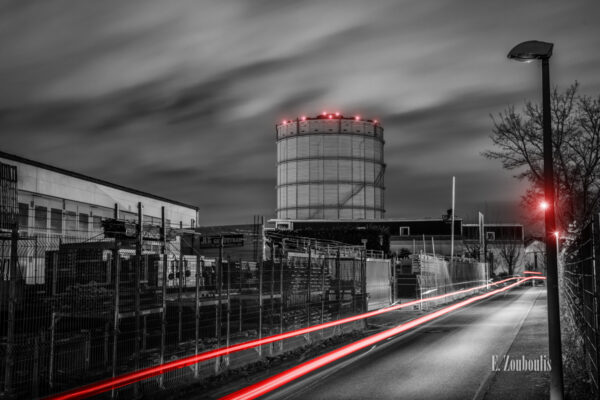 Schwarzweiß Aufnahme bei Nacht am Gaskessel in Stuttgart Ost (Gaisburg). Im Vordergrund rote Lichtschweife, die den vorbeifahrenden Verkehr sichtbar machen. Der Gasturm ist hinter einer Industrieanlage mit roten Lichteffekten zu sehen. Über dem Gasturm ziehen die Wolken vorbei, die durch das Licht Stuttgart erhellt werden