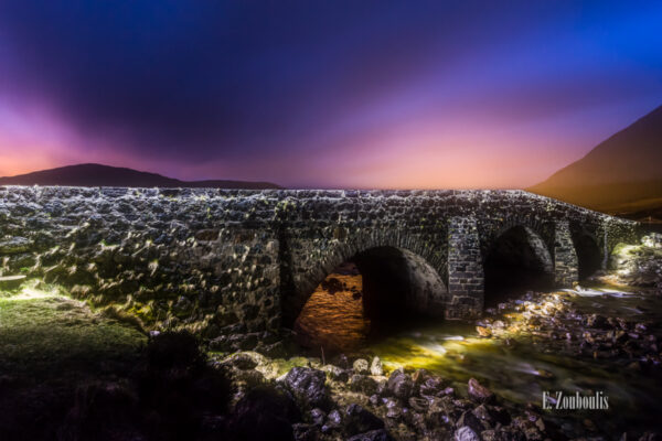 Nachtaufnahme an der Sligachan Bridge auf der Isle of Skye, Schottland. im Vordergrund die Felsen und das Wasser, das unter der Brücke hindurchfließt. In der Mitte des Bildes ist die Brücke zu sehen. Im Hintergrund die Lichter der Nacht und die Silhouetten der Berge