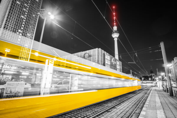 Schwarzweiss-Aufnahme einer einfahrenden Straßenbahn am Alexanderplatz in Berlin. Schwarzweiss mit farbigem Lichtschweif, der die Straßenbahn erkennen lässt