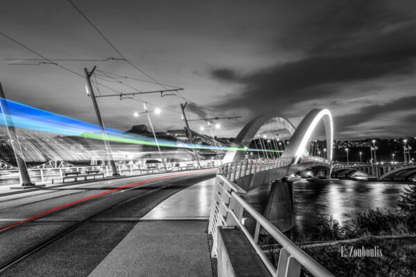 Schwarzweiss-Aufnahme an der Pont Raymond Barre Brücke in Lyon, Frankreich. Im Vordergrund zieht eine Bahn über die Brücke und hinterlässt einen farbigen Lichtschweif. Der Fluss Rotten ist rechts im Bild zu sehen und im Hintergrund befindet sich das Musée des Confluences.