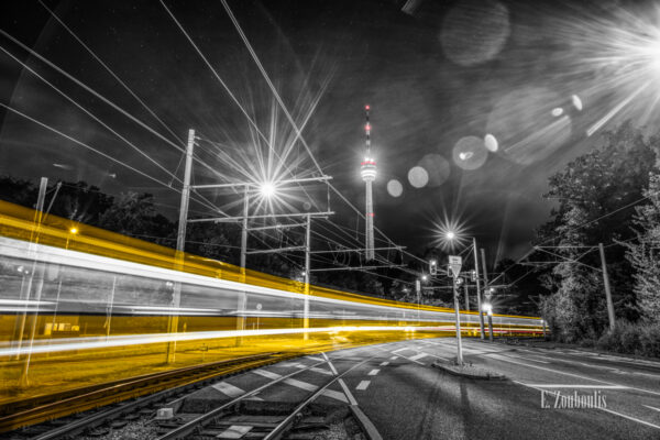 Schwarzweiß-Aufnahme mit roten und gelben Elementen mit Blick auf den Fernsehturm in Stuttgart Degerloch. Eine Bahn fährt am Betrachter vorbei und hinterlässt einen gelben Lichtschweif