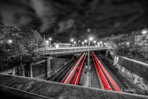 Schwarzweiss-Aufnahme an der Jurastraße in Stuttgart Vaihingen. Im Vordergrund sind vorbeifahrende Bahnen zu sehen, die einen roten Lichtschweif hinterlassen. In der Mitte des Bildes sieht man die Haltestelle Jurastraße und im Hintergrund ist der Vaihinger Colorado Turm zu sehen