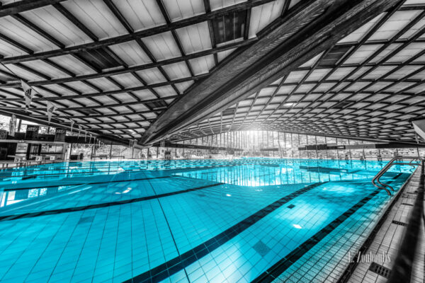 Schwarzweiß Aufnahme im Inneren des Badezentrum Sindelfingen. Das Wasser in den Becken ist in blau zu sehen. Seitliche Ansicht auf das 50m Becken. Im Hintergrund ist der Sprungturm zu sehen