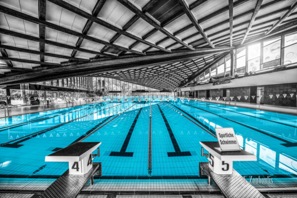 Schwarzweiß Aufnahme im Inneren des Badezentrum Sindelfingen. Das Wasser in den Becken ist in blau zu sehen. Frontal-Ansicht auf das 50m Becken. Im Hintergrund ist der Sprungturm zu sehen