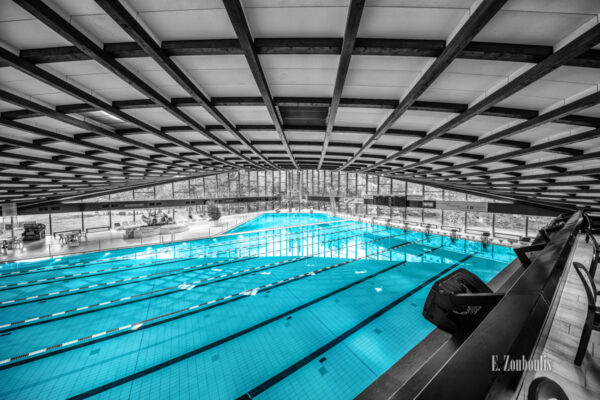 Schwarzweiß Aufnahme im Inneren des Badezentrum Sindelfingen. Das Wasser in den Becken ist in blau zu sehen. Seitliche Ansicht von oben auf das 50m Becken. Im Hintergrund ist der Sprungturm zu sehen