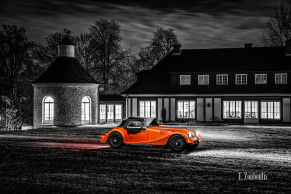 Schwarzweißaufnahme eines Orangenen Morgan 4/4. Fahrzeug in Orange in der Mitte des Bildes. Im Hintergrund ist die Villa Schwalbenhof in Gärtringen zu sehen