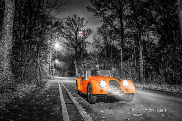 Schwarzweißaufnahme eines orangenen Morgan 4/4 Cabrio mit offenem Verdeck. Fahrzeug in Orange in der Mitte des Bildes. Im Hintergrund ist der Fernsehturm in Stuttgart Degerloch zu sehen