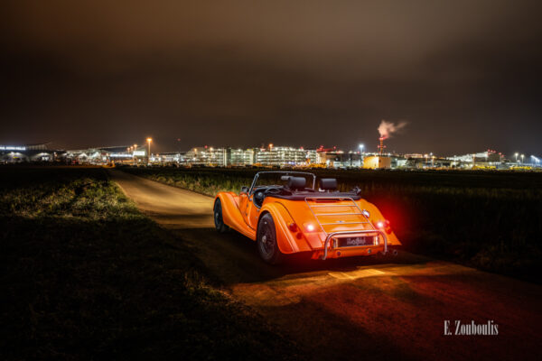 Aufnahme eines orangenen Morgan 4/4. Fahrzeug in Orange in der Mitte des Bildes. Im Hintergrund ist die Messe Stuttgart in der Nähe vom Flughafen Stuttgart zu sehen