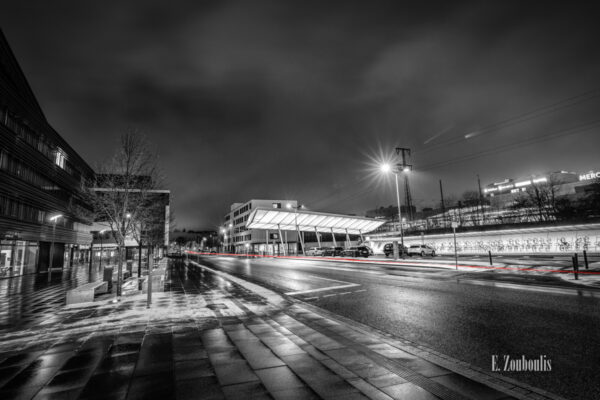 Schwarzweiß-Aufnahme am Bahnhof in Böblingen. Im Vordergrund liegt ein wenig Schnee, die Straße und der Gehweg sind nass. Auf der Straße Fähr ein Auto vorbei und hinterlässt einen roten Lichtschweif. In der Mitte des Bildes sieht man die Dachkonstruktion, die zu den Gleisen führt