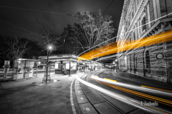 Schwarzweiß-Aufnahme einer Straßenbahn mit gelbem Lichtschweif vor einem Würstelstand am Burgring in Wien