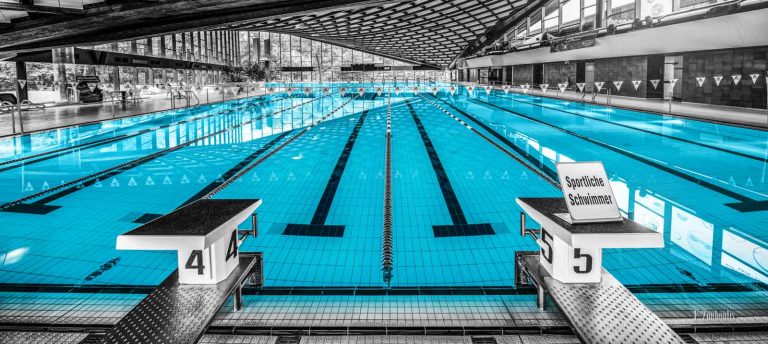 Schwarzweiß Aufnahme im Inneren des Badezentrum Sindelfingen. Das Wasser in den Becken ist in blau zu sehen. Frontal-Ansicht auf das 50m Becken. Im Hintergrund ist der Sprungturm zu sehen