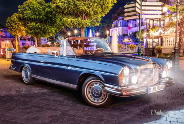 Ein 1971 Mercedes-Benz 280 SE nachts im Europa-Park seitlich von vorne mit den Lichtern des Europapark im Hintergrund