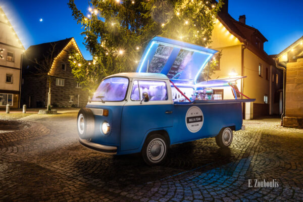 Wandbild eines Volkswagen Bulli T2 aus dem Jahr 1970. Der Oldtimer ist als Bar umgebaut worden und leuchtet vor einem Weihnachtsbaum