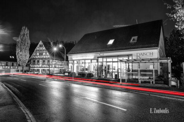 Nachtaufnahme in Aidlingen bei Böblingen. Zu sehen ist die Hauptstraße und die Bäckerei Raisch. Davor sind rote Lichtschweife, die den Verkehr kennzeichnen