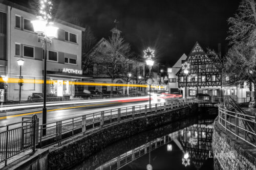 Nachtaufnahme in Aidlingen bei Böblingen. Zu sehen ist die Hauptstraße und die Aid, in der sich das Fachwerkhaus der Metzgerei Zweygart spiegelt. Entlang der Straße sind rote und gelbe Lichtschweife, die den Verkehr kennzeichnen.