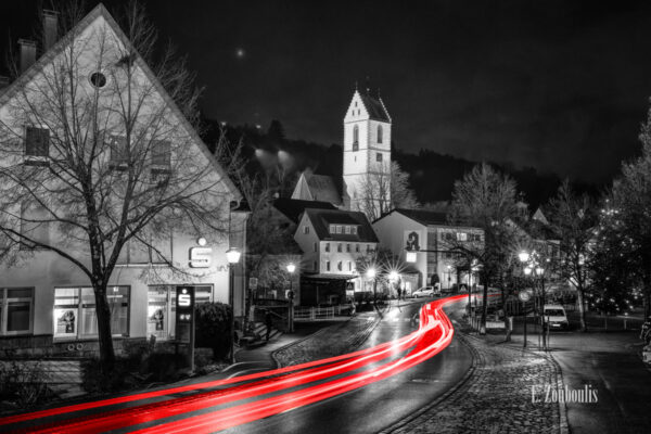 Nachtaufnahme in Aidlingen bei Böblingen. Zu sehen ist die Hauptstraße und die Sparkasse. Davor sind rote Lichtschweife, die den Verkehr kennzeichnen. Im Hintergrund steigt der Kirchenturm der evangelischen Kirchengemeinde empor