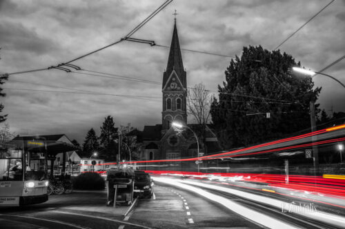 Nachtaufnahme am Quirinusplatz in Bonn Dottendorf mit Blick auf die St Quirinuskirche bei Nacht. Zu sehen ist neben der Kirche und den Lichtschweifen des vorbeiziehenden Verkehrs, eine Straßenbahn