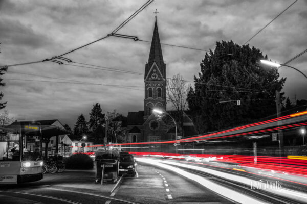 Nachtaufnahme am Quirinusplatz in Bonn Dottendorf mit Blick auf die St Quirinuskirche bei Nacht. Zu sehen ist neben der Kirche und den Lichtschweifen des vorbeiziehenden Verkehrs, eine Straßenbahn